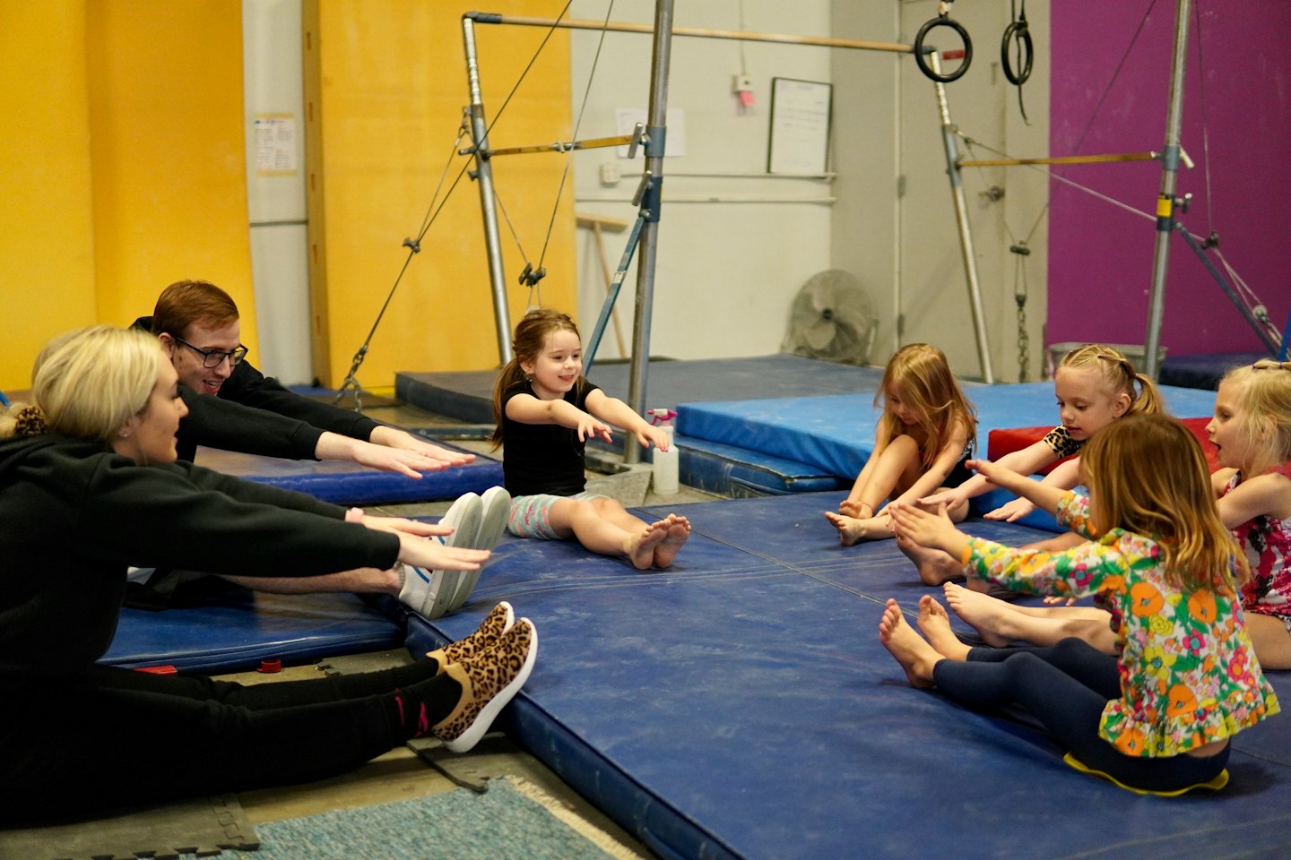 Children and adults stretching on blue mats in a gymnastics studio.