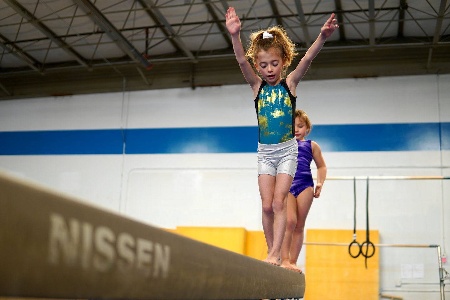 Two girls practice gymnastics on a beam in a gym.