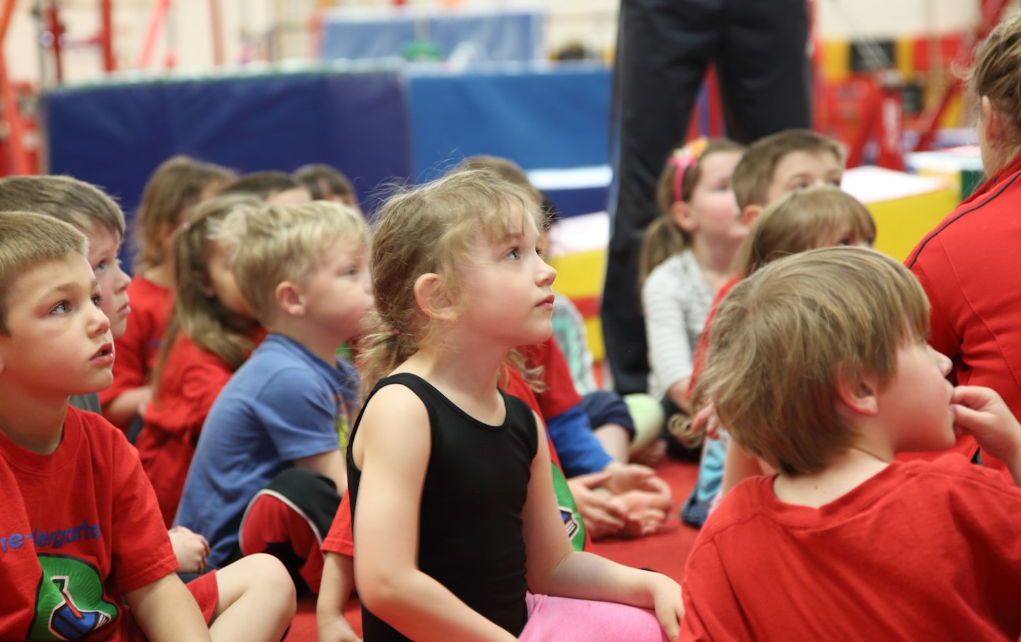 Children sit on a red mat in a gym looking up.