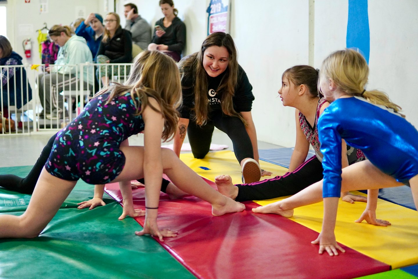 Gymnastics coach kneels and smiles at young girls stretching.
