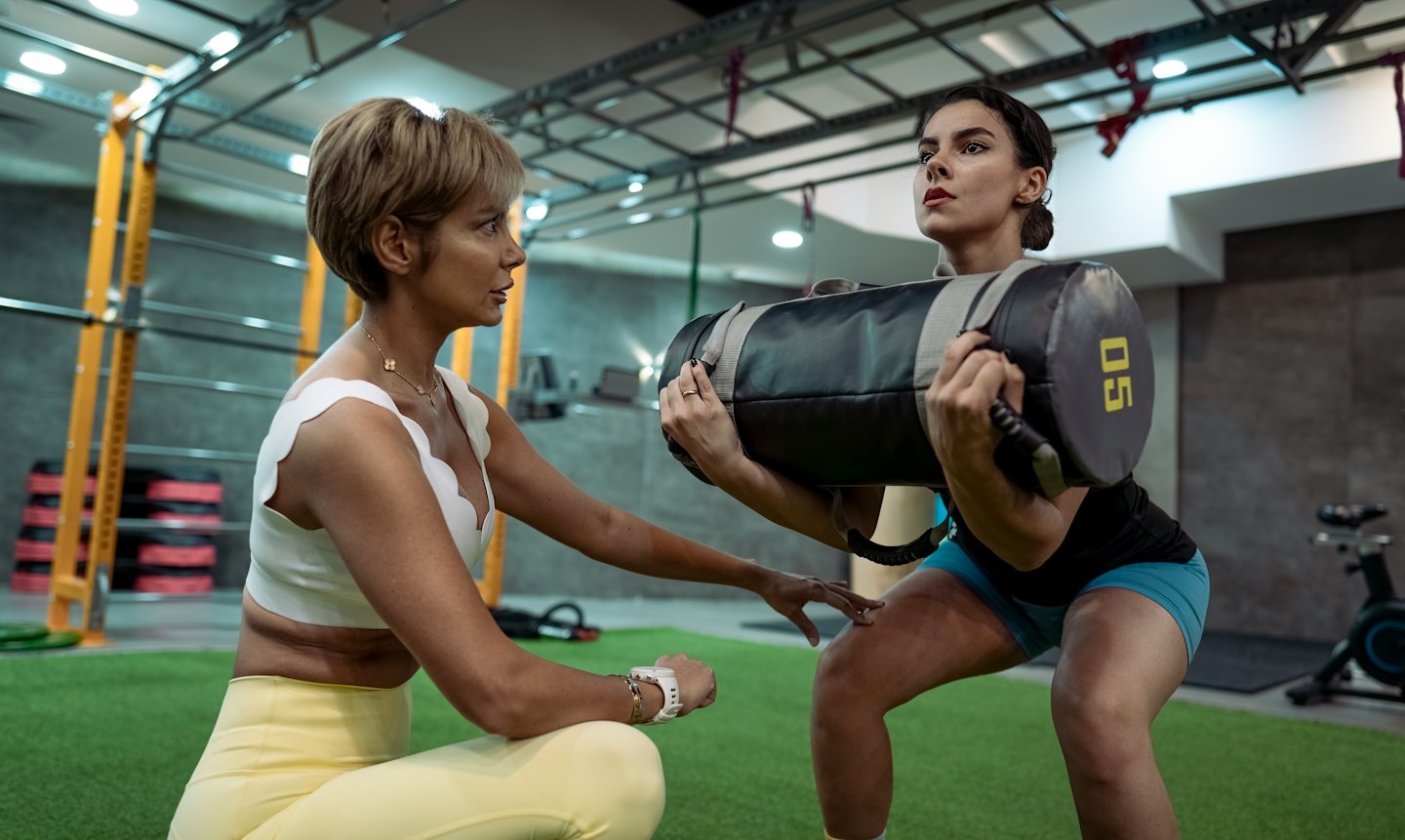Coach guiding woman performing squats with sandbag in gym.