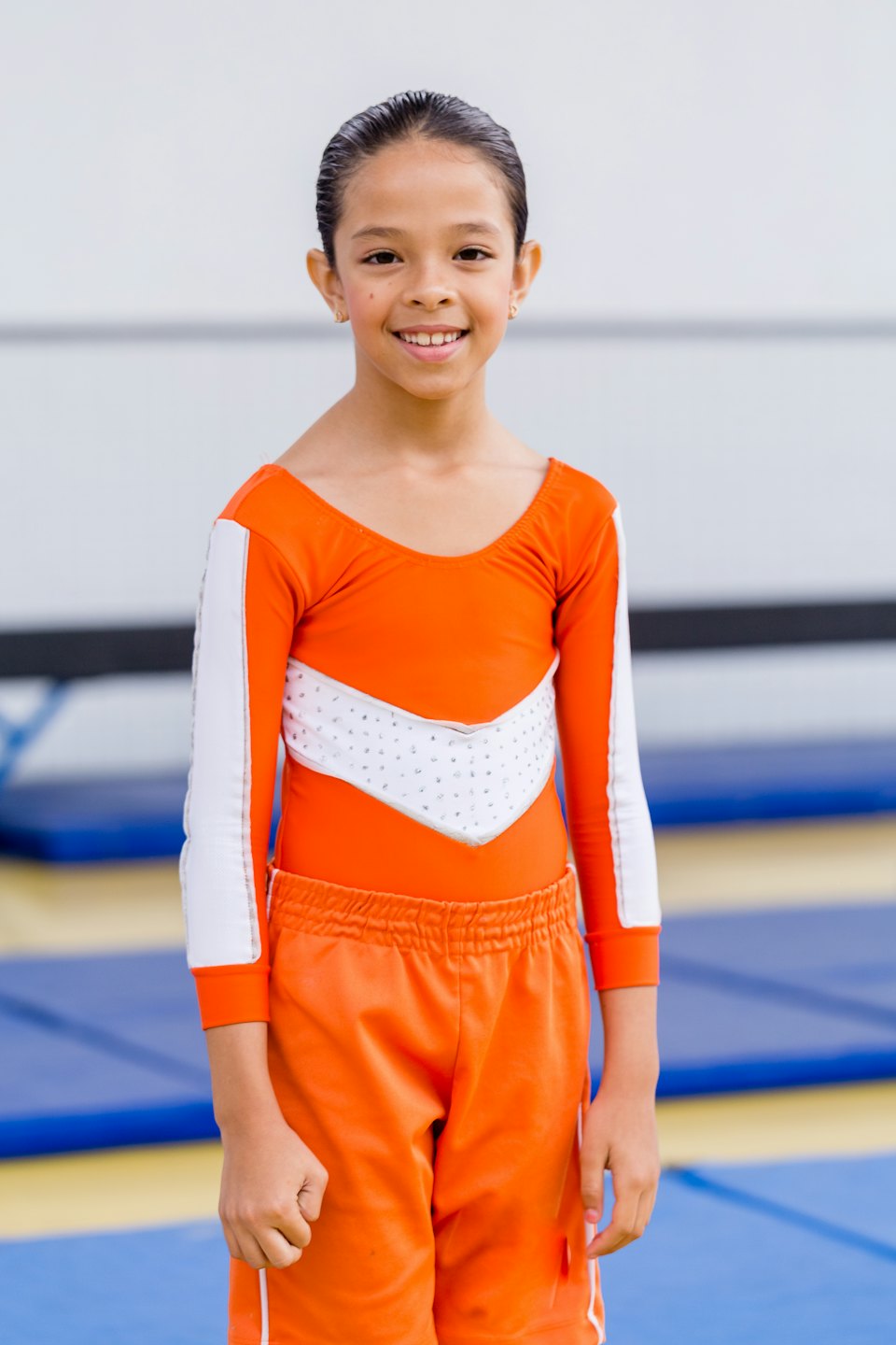 Gymnast in an orange leotard stands on a gym mat.