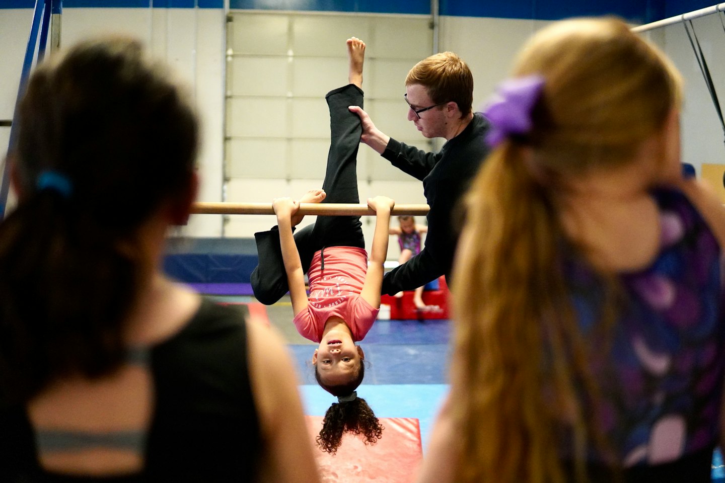 Gymnastics instructor helps a girl hanging upside down on a bar.