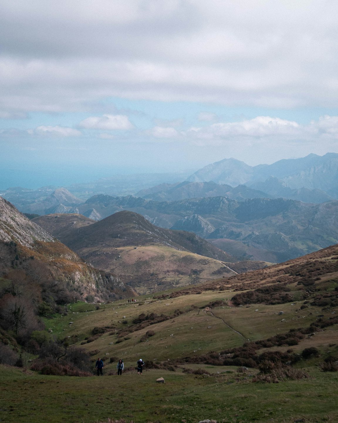 Hikers on a mountain path with distant ocean under cloudy sky.